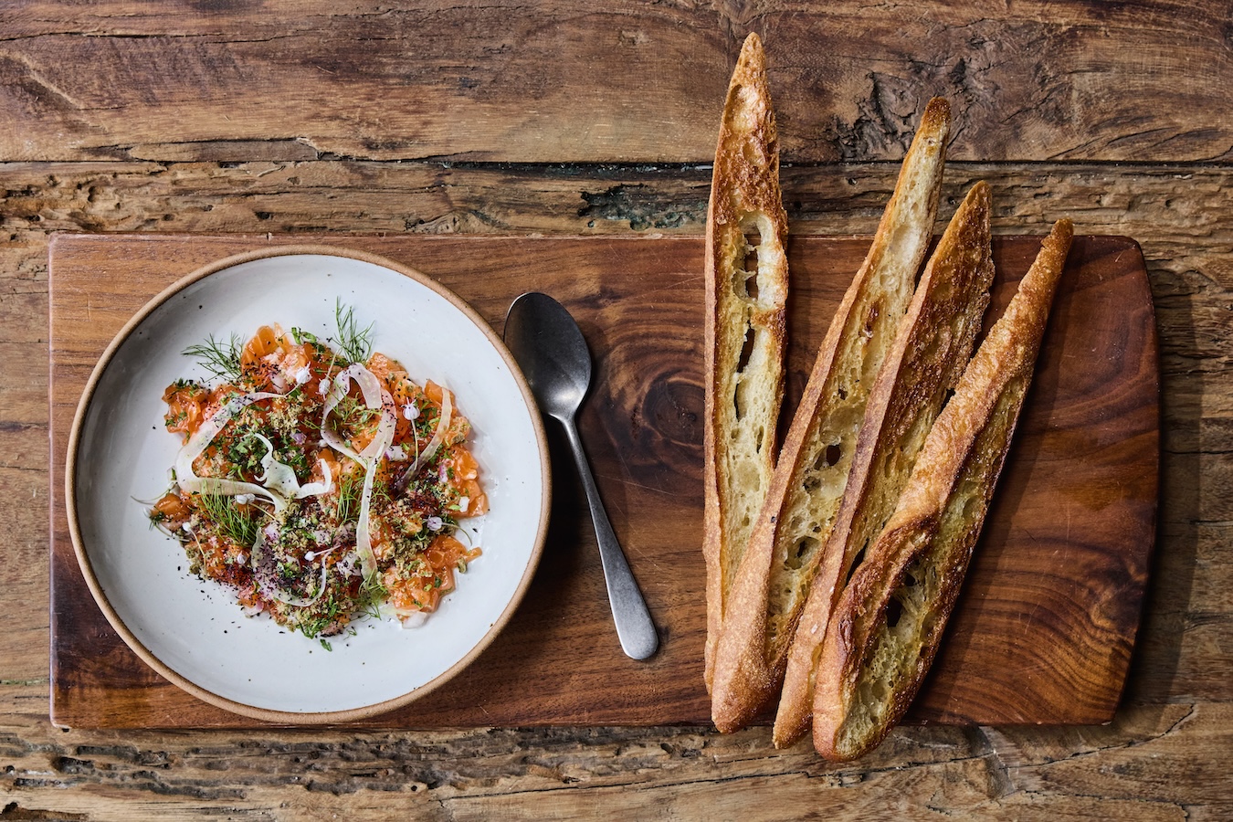 A white plate on a wooden table with trout tartar garnished with fennel. A wooden plank with toasted bread sits next to it.