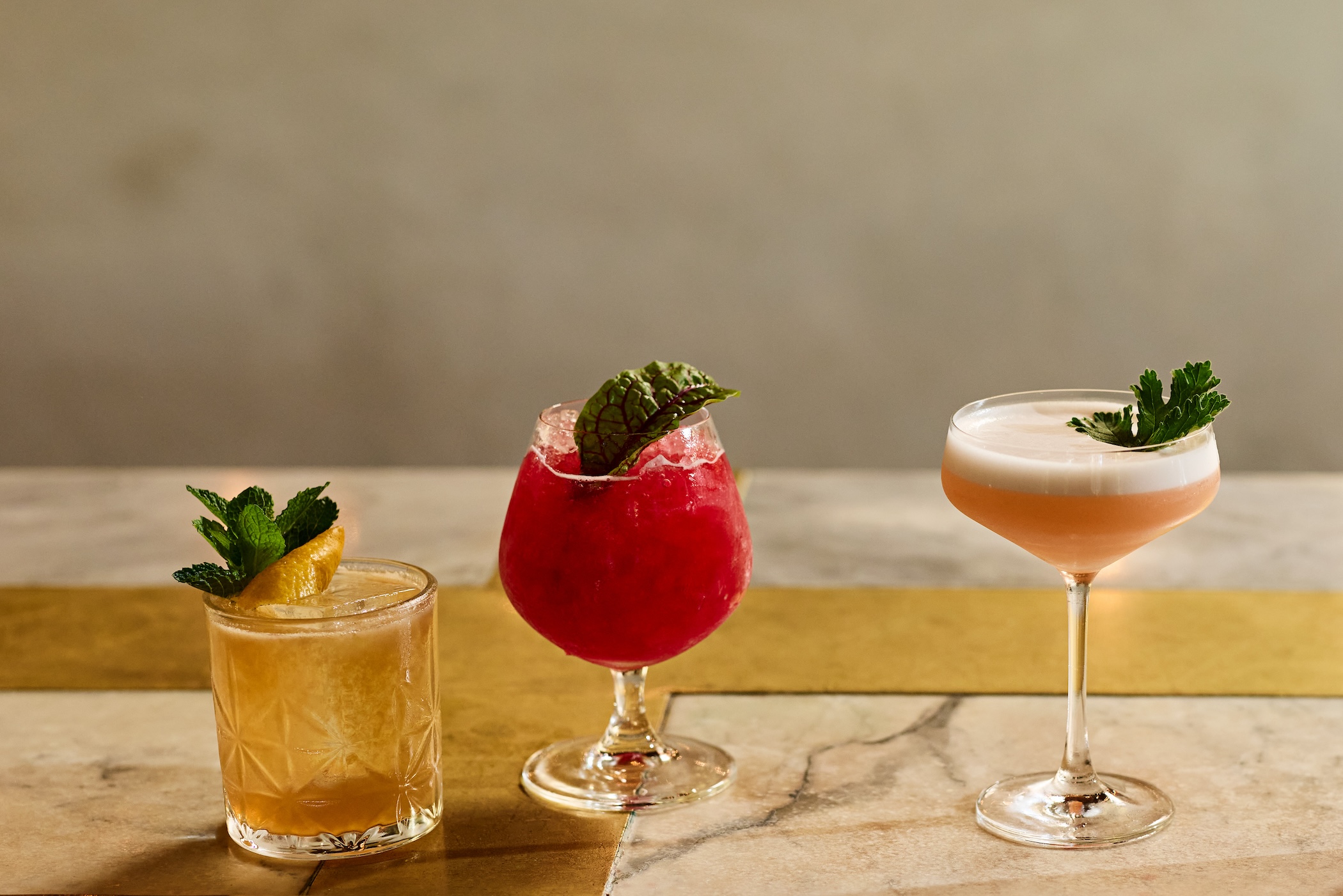 Three cocktails in a line on a bar in different glasses, all garnished with herbs.
