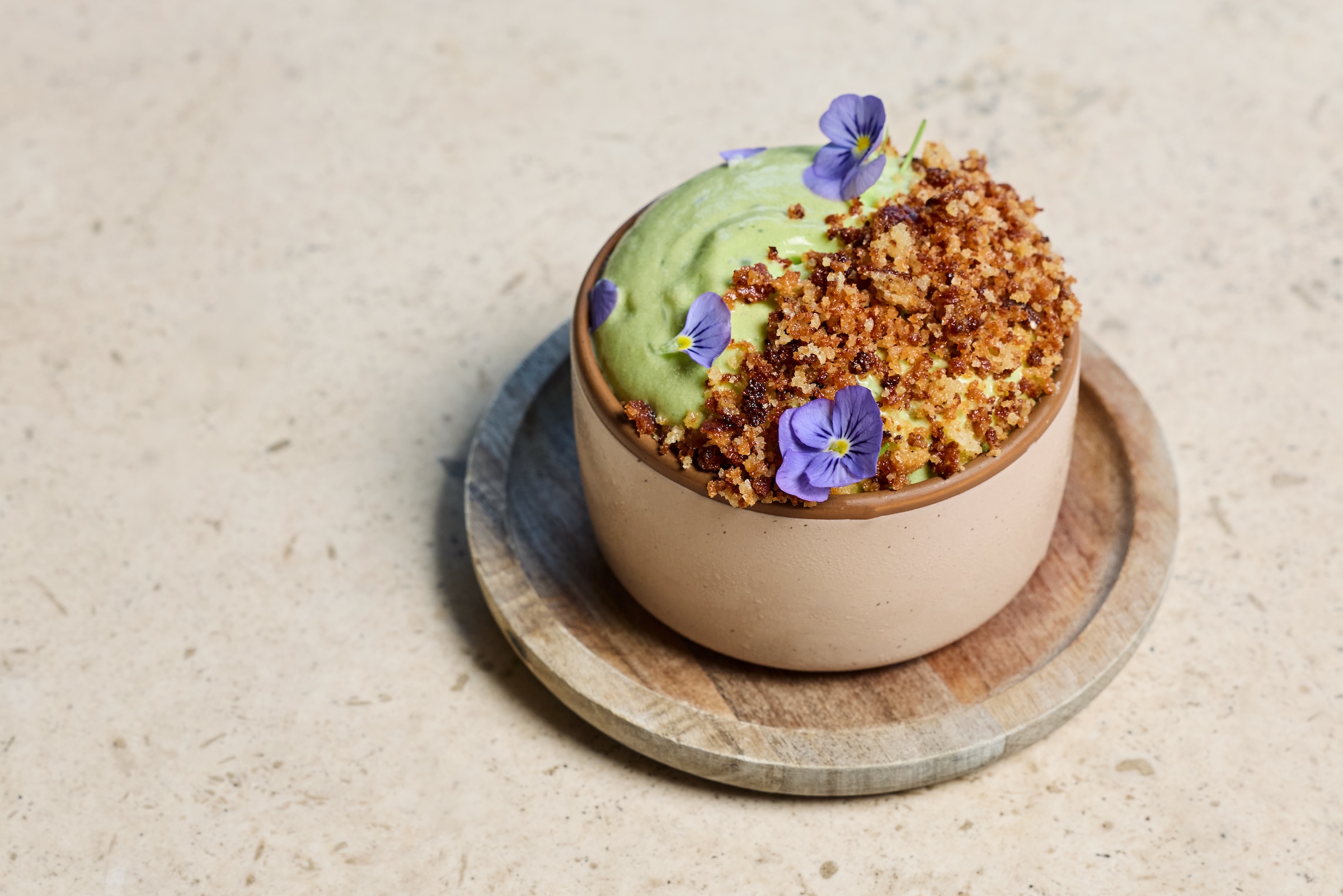 A small ceramic dessert pot full of green basil tres leches. Topped with green whipped cream, purple flowers, and a crumble.