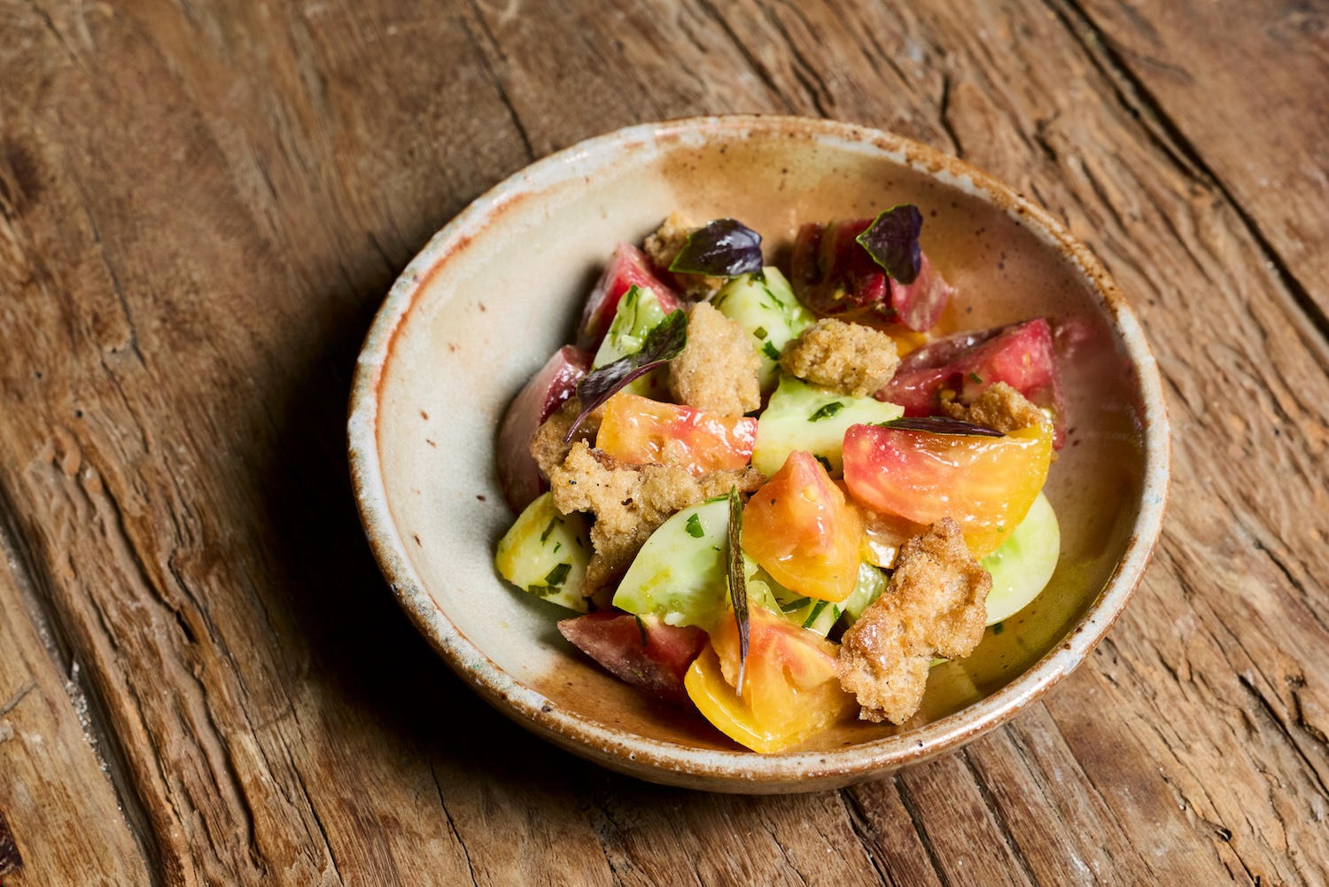 a textured bowl full of orange and red heirloom tomatoes, cucumbers, and bread peices.