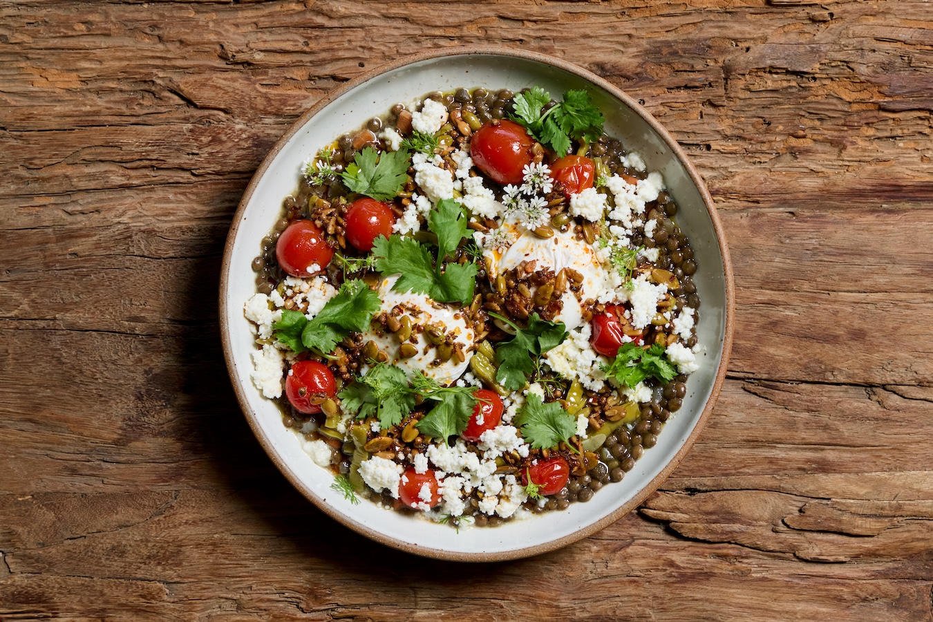 A white bowl full of bowl lentils , cherry tomatoes, crumbled white cheese, cilantro and white flower blossoms.