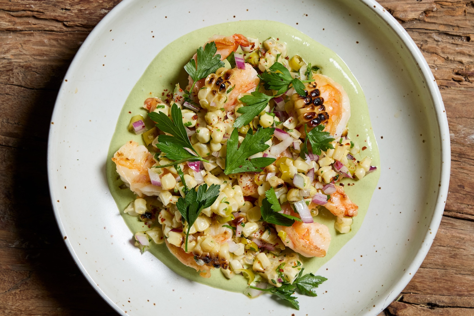 A white bowl full of grilled corn and shrimp over a green sauce and topped with fresh green herbs.