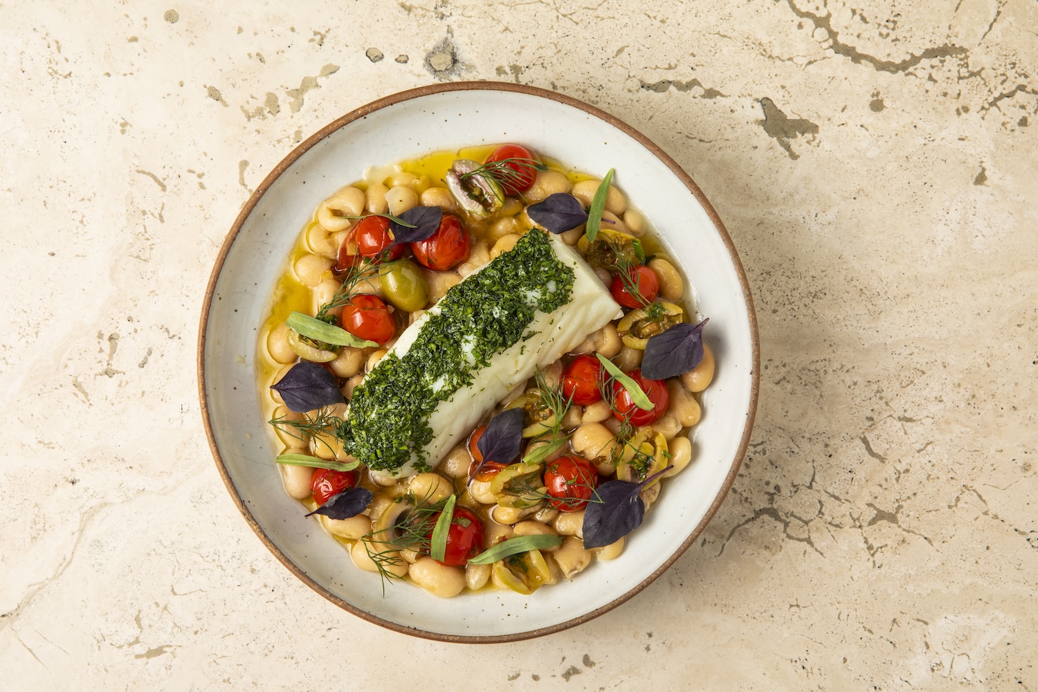 A white fish covered in a green herb pate sitting on a bed of white beans, tomatoes and olives on a white plate.