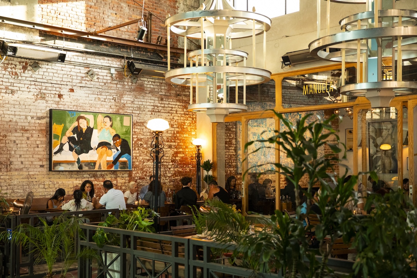 Main dining room at Manuela. People are seated and eating next to a brick wall and green plants.