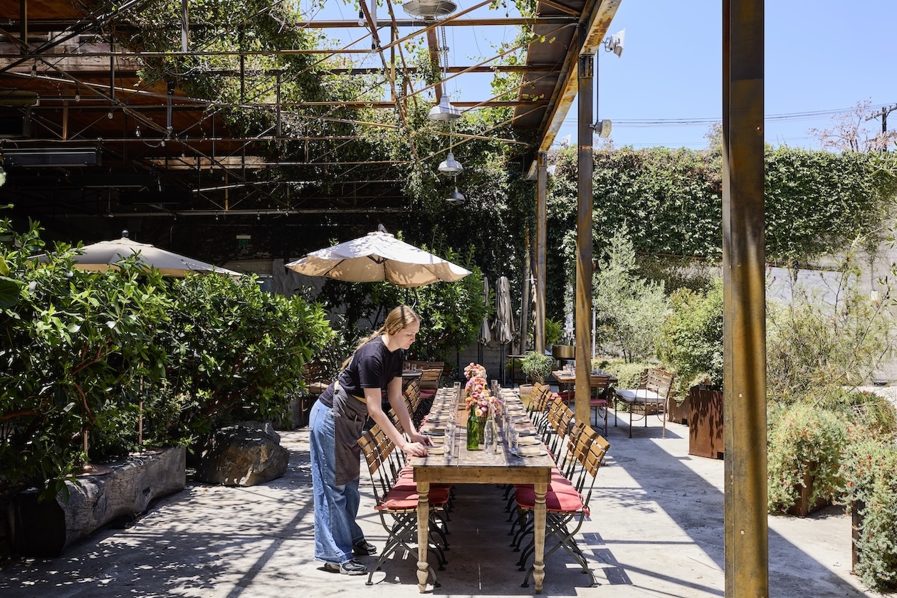 Outdoor dining setup with a long wooden table being prepared under a pergola covered in greenery, surrounded by plants and patio umbrellas in a sunny courtyard garden.