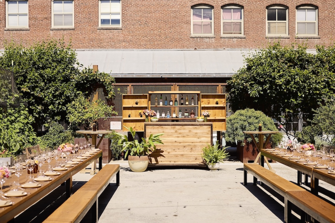 Two long tables set for dinner service in an outdoor garden area with a wooden bar framed in between them towards the back of the space.