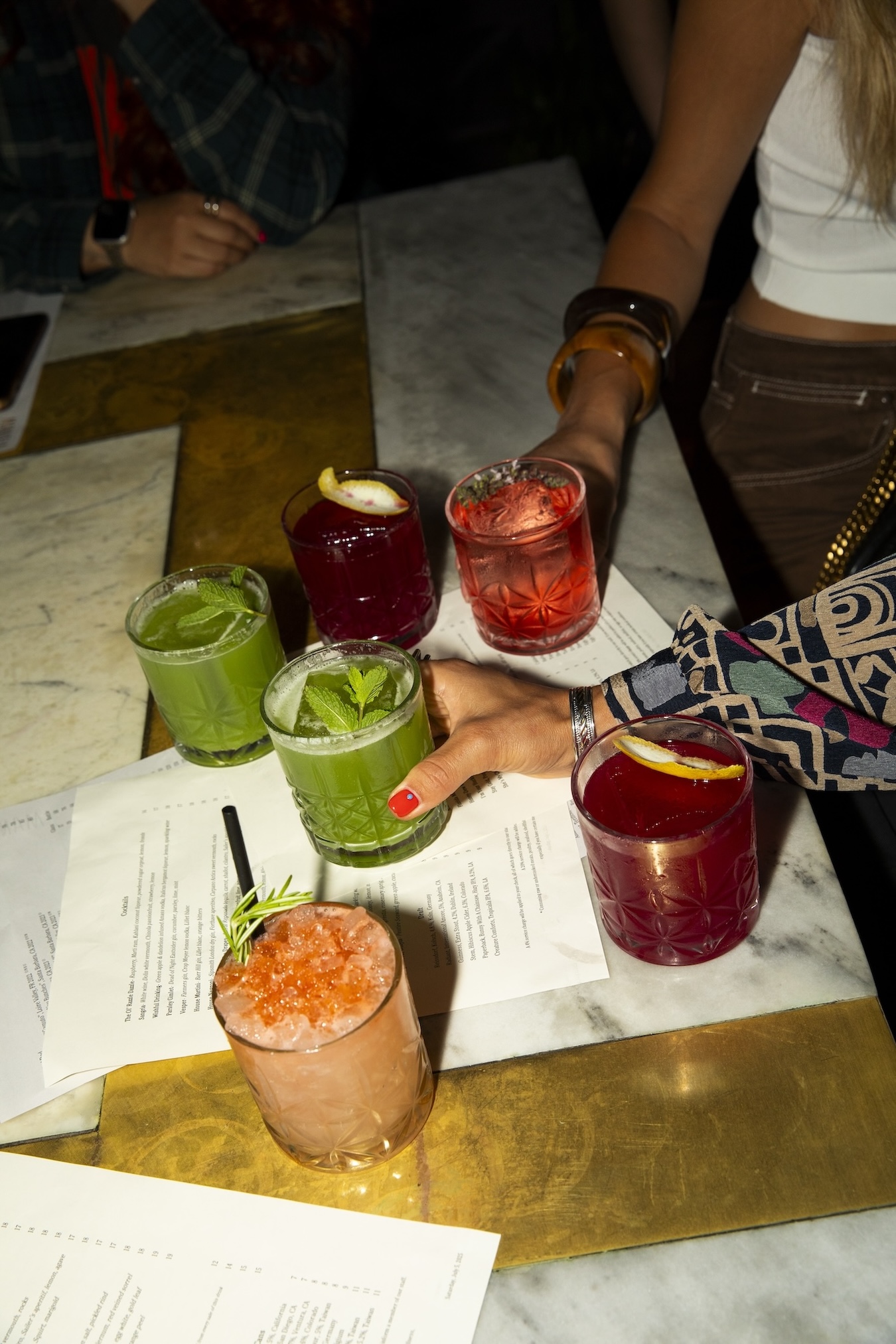 A collection of green, purple, and peach colored cocktails on a table with two separate hands holding two of the cocktails.