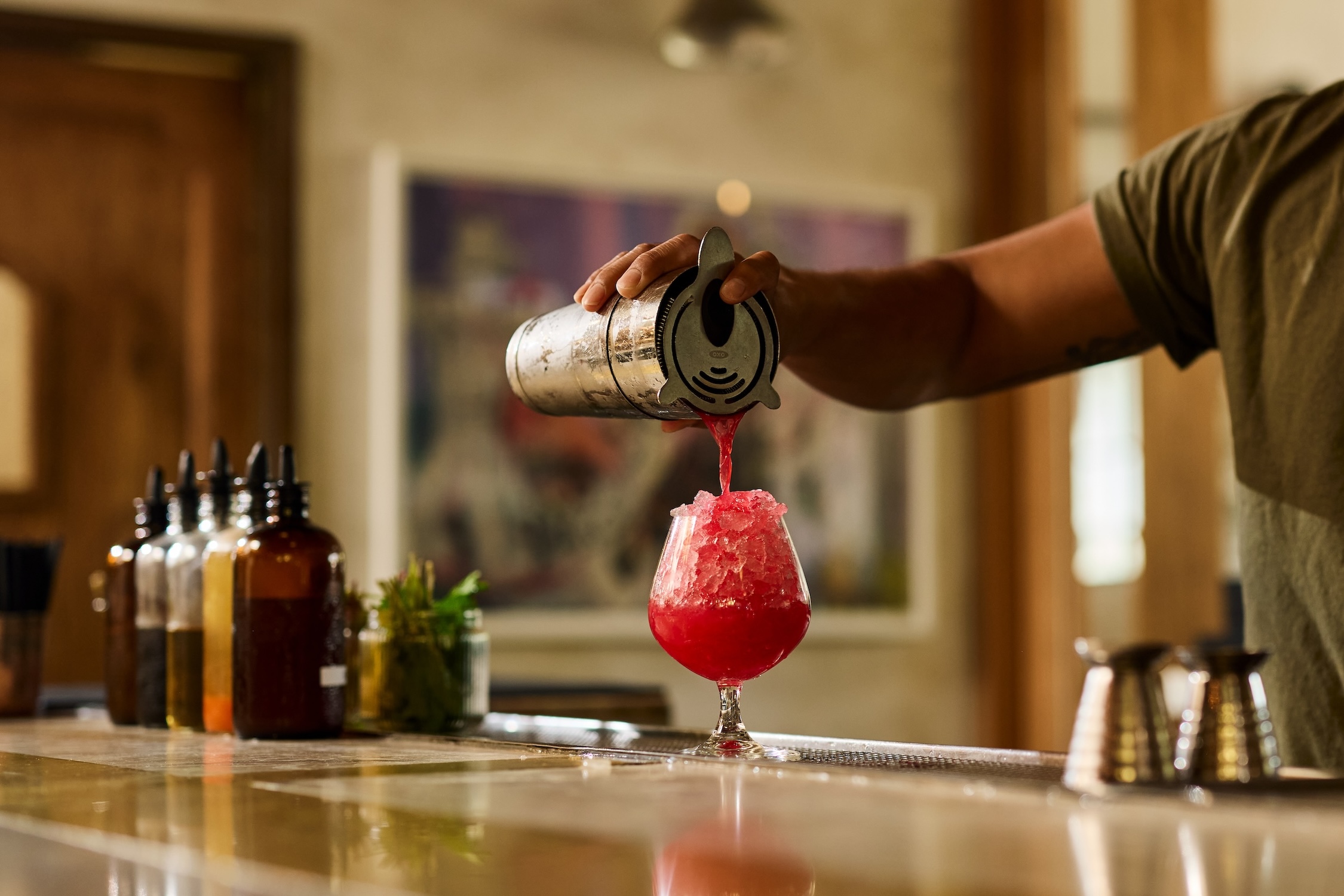A hand holding a mixing shaker, pour a fresh bright red cocktail into a glass over pebble ice.