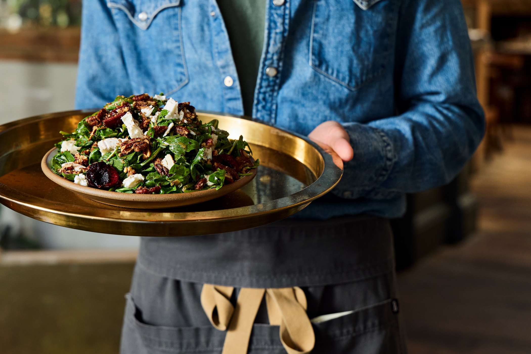 A server holding a tray with a plate piled high with a salad of arugula, nuts, white cheese, and more.