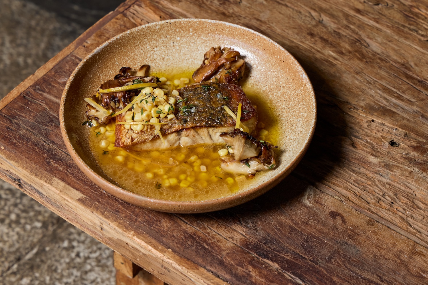 A shallow brown bowl with a cooked piece of fish, skin side up, in a clear broth with mushrooms and corn.