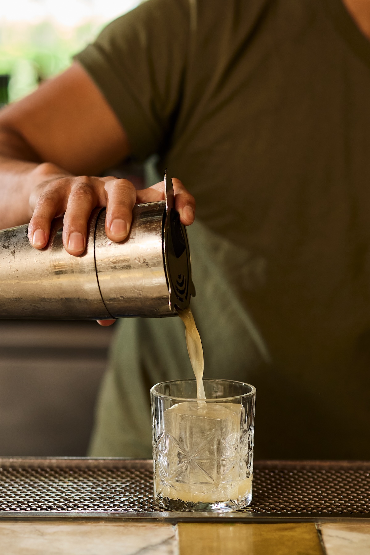 A hand holding a mixing shaker, pour a fresh cocktail into a rocks glass over a big piece of ice
