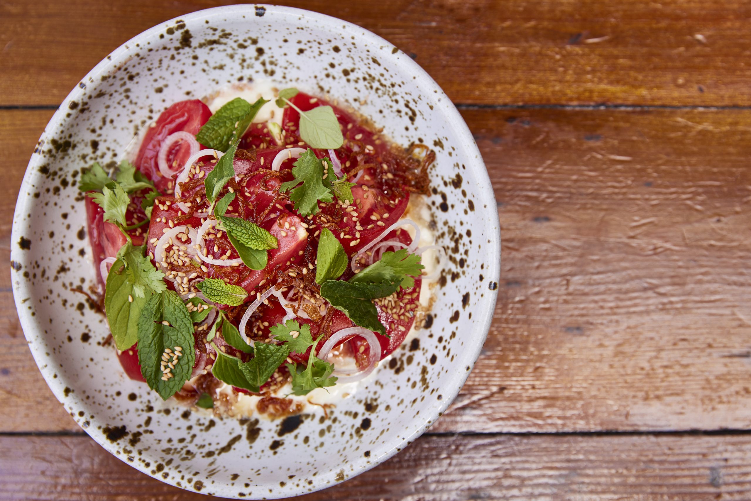Textured white and brown speckled bowl on a wooden surface. Bowl is full of read tomatoes topped with green herbs