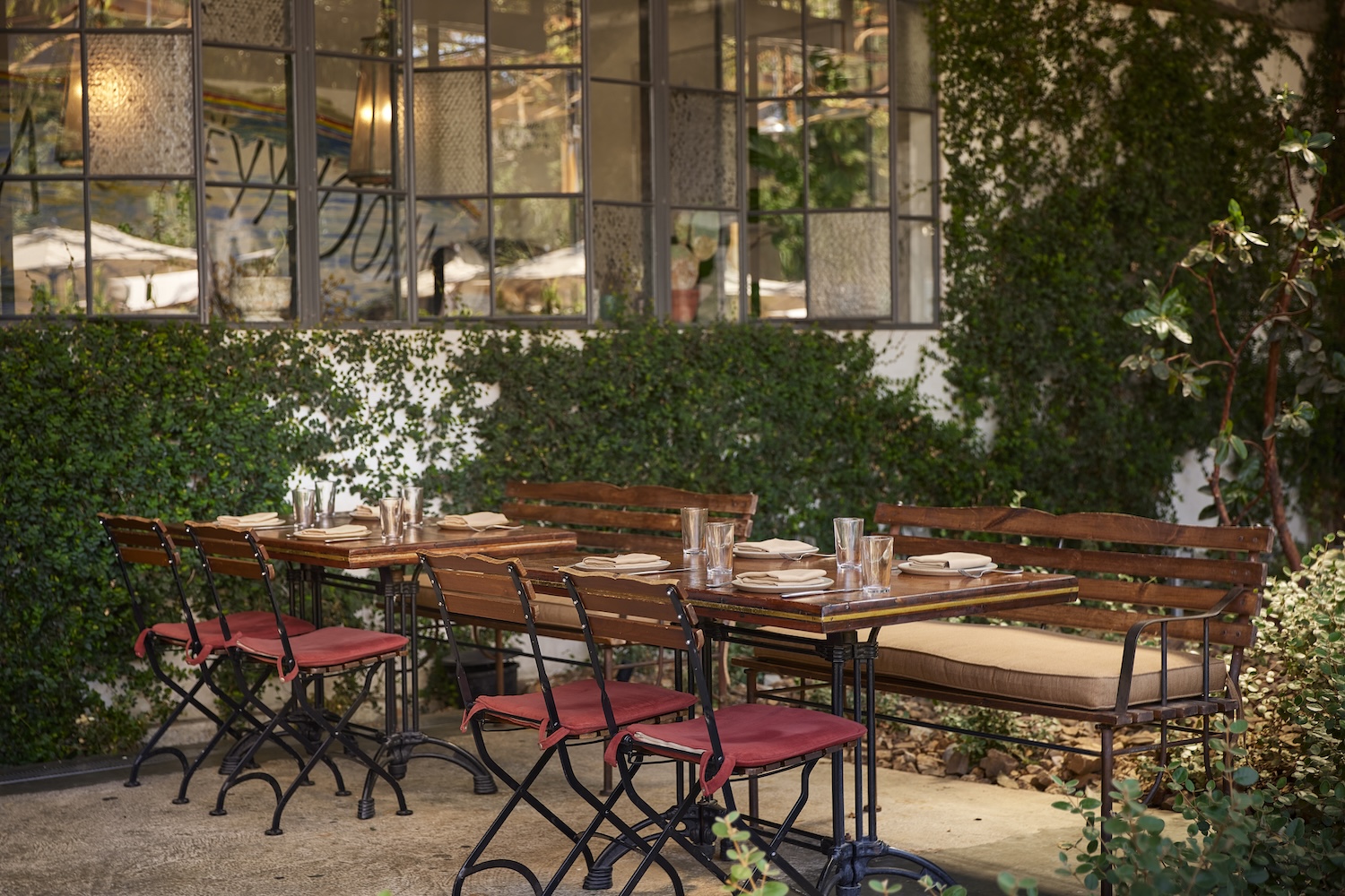 A rustic outdoor dining area with wooden tables and chairs, cushioned seating, and greenery covering the surrounding walls.