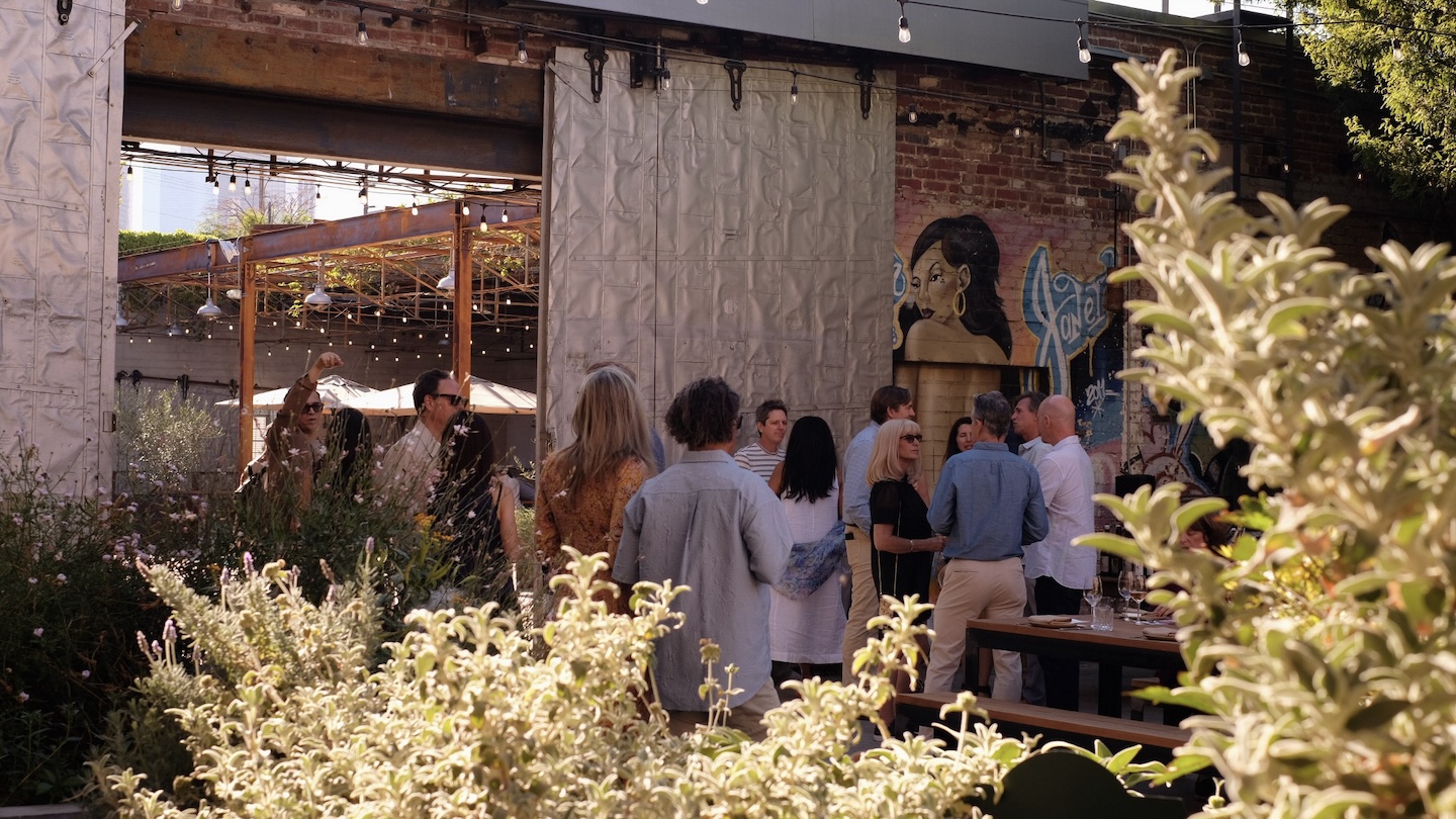 Holiday Wine Dinner at Manuela. People standing outdoors in front of a large metal door, with light green and dark green bushes in front of them.