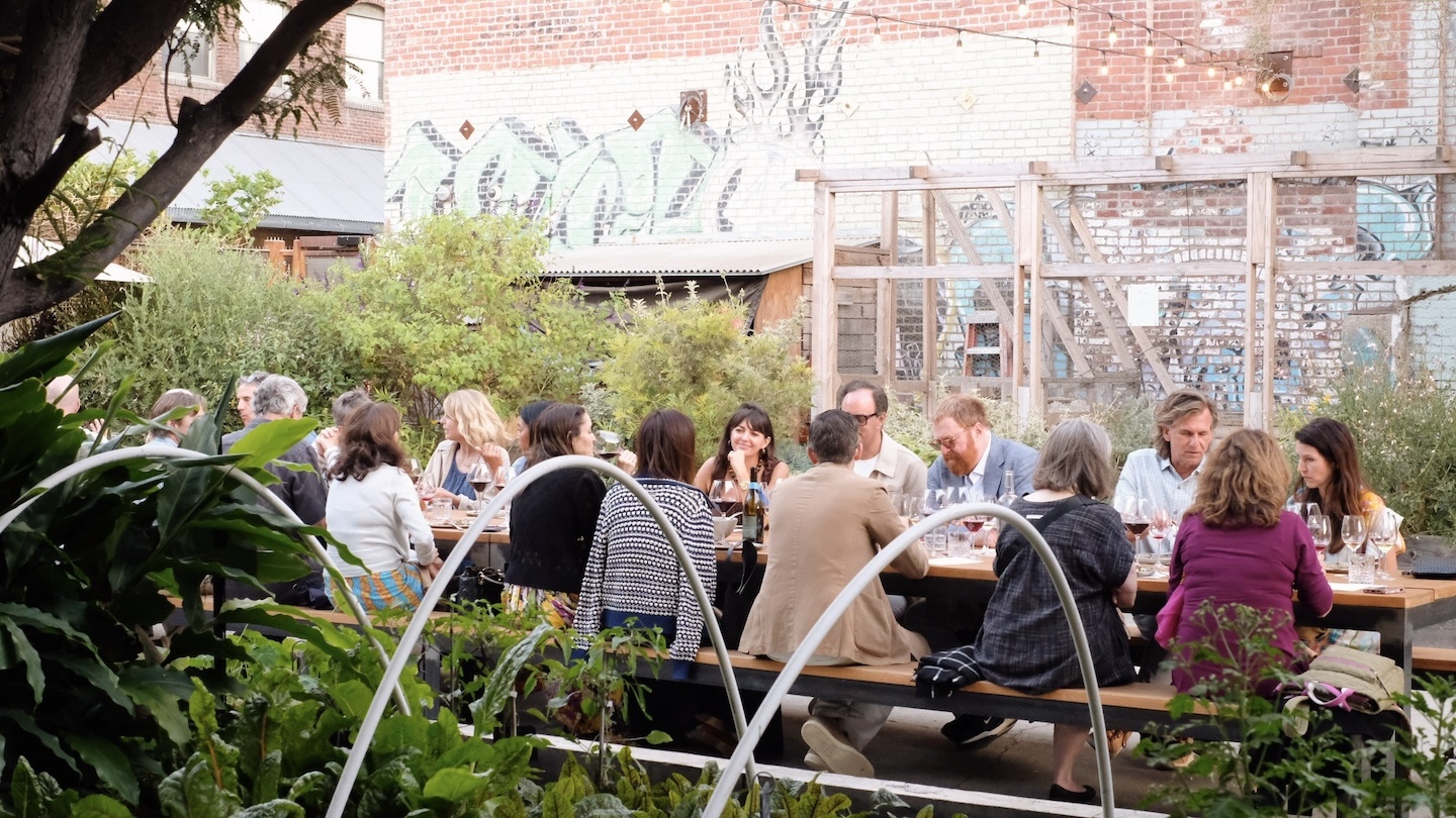 People sitting at a long table outside enjoying a meal together for the Holiday Wine Dinner at Manuela. The are surrounded by light green bushes, a small garden and a large chicken coop.