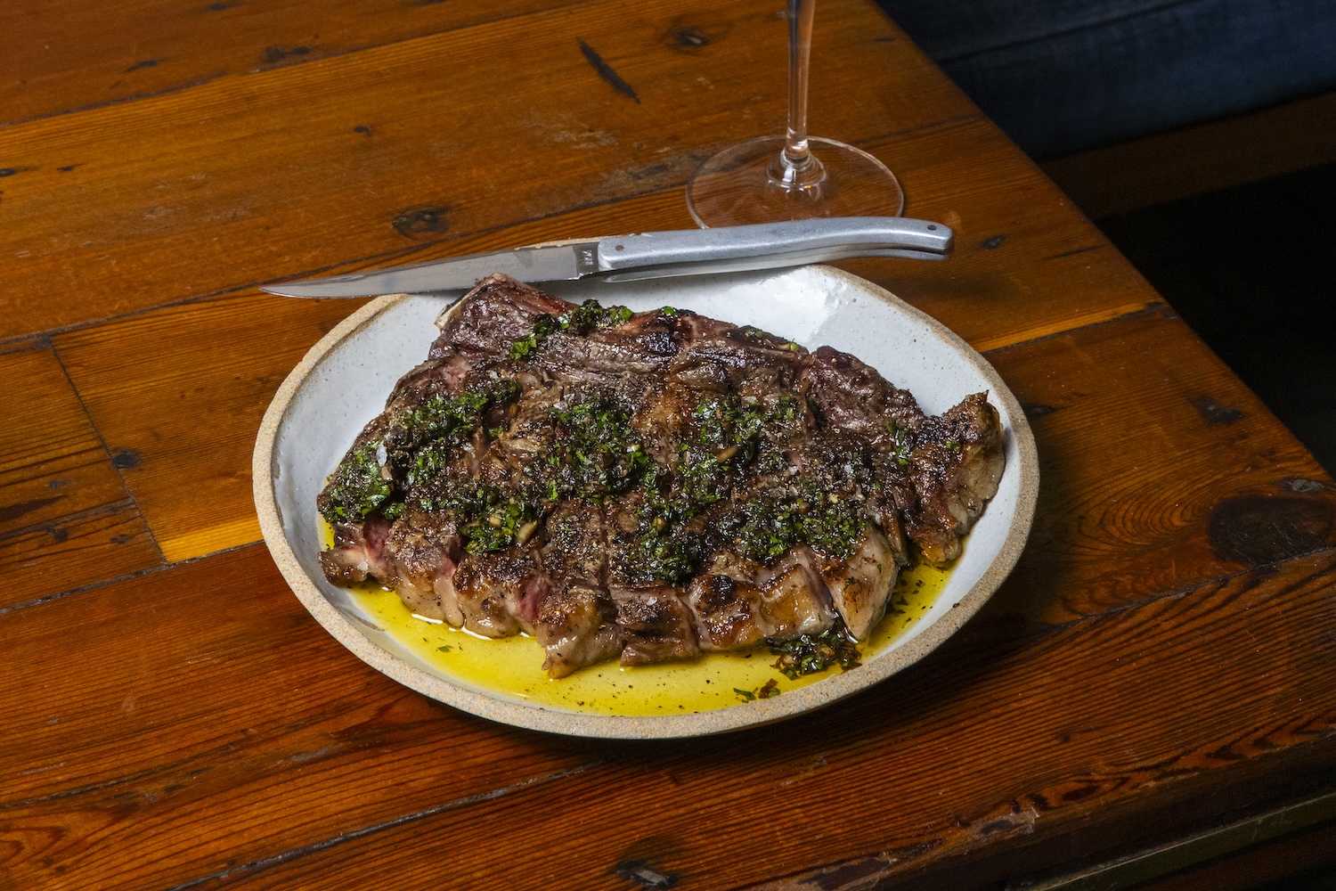 A sliced steak on a white plate covered in dark green chimichurri sauce. The dish is the Double R Ranch bone-in 18oz ribeye plated with a steak knife.